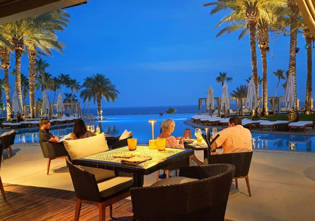 Tables with chairs along the infinity pool with palm trees and the sea in the distance at Vela in the Hilton Los Cabos.