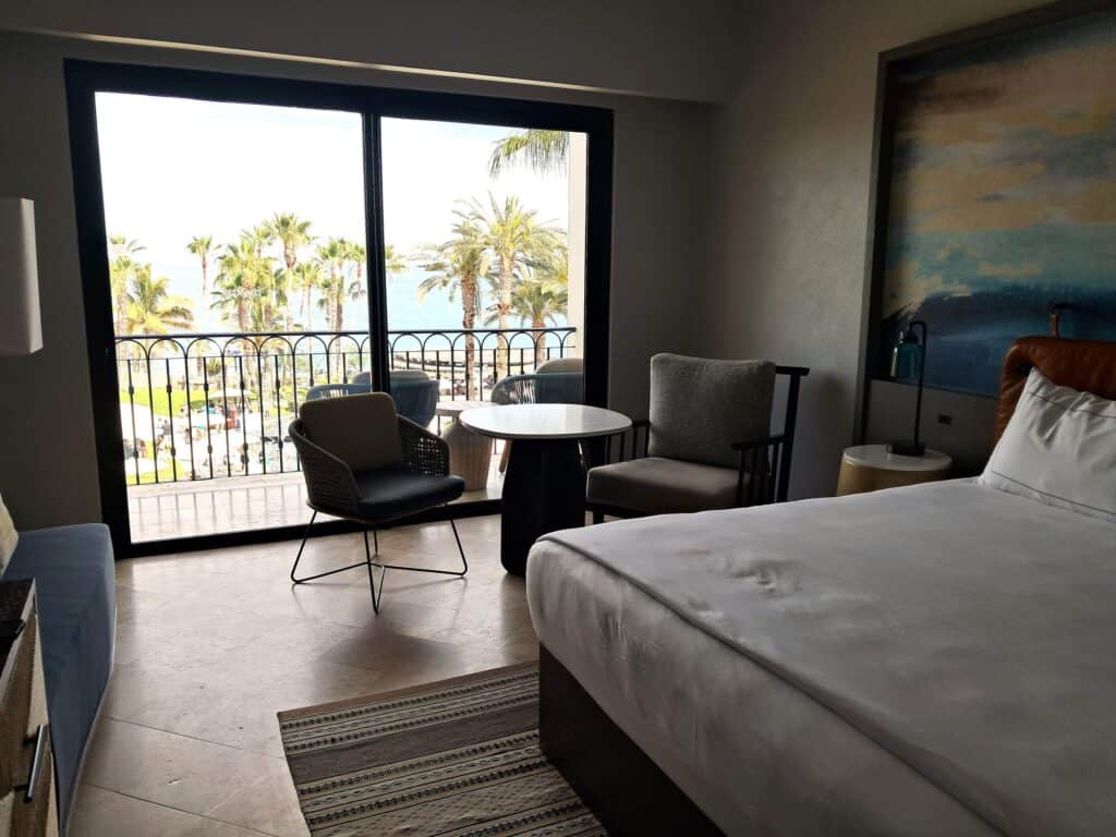 Studio room with bed, 2 chairs, table and sliding glass doors to the balcony.