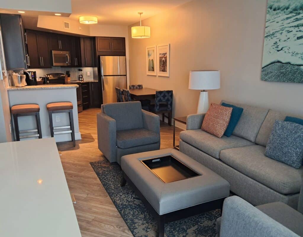Living room with sofa, two chairs, and kitchen in background with a table and chairs, bar with 2 stools, fridge, oven and cabinets in the Ocean 22 2-bedroom unit.