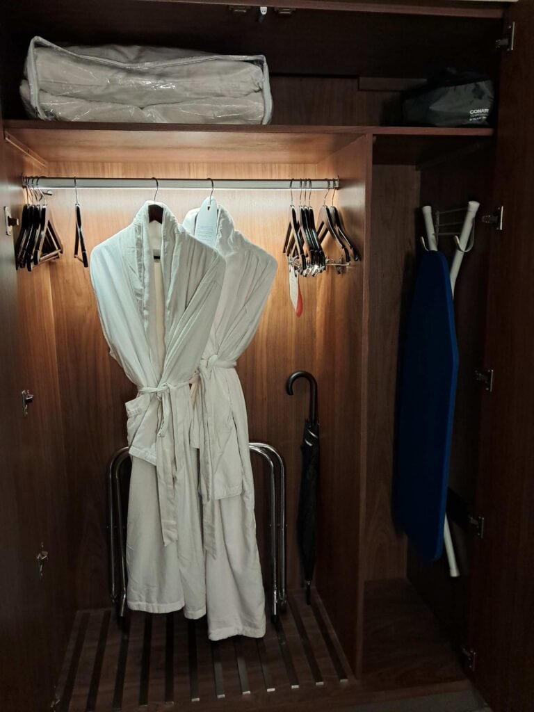 Two white robes hanging in the closet with an umbrella and an ironing table in the Residences by Hilton Club review studio room.
