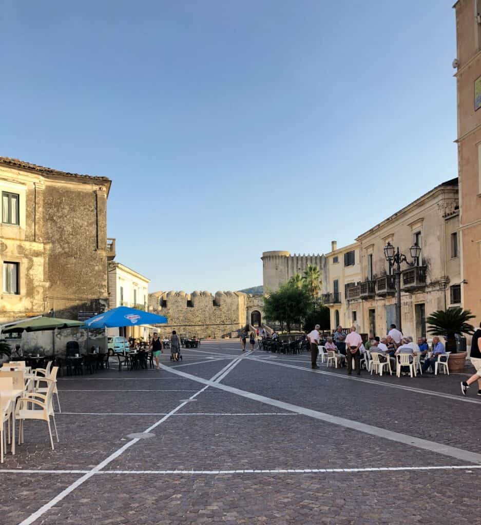 Piazza Campo in the center of Santa Severina with tables and chairs for afternoon drinks surrounded by historic building and a castle in the background in Santa Severina.