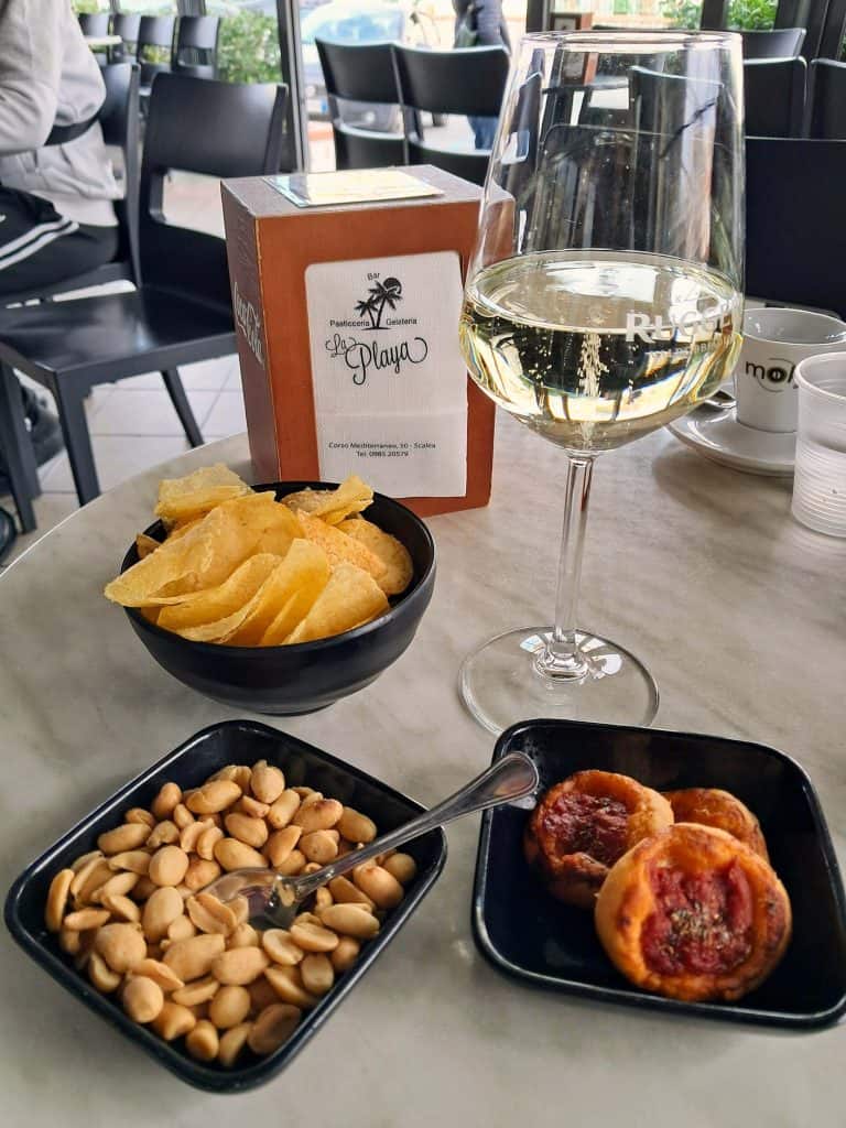 A glass of prosecco with a bowl of potatoes chips, nuts, and small pizza snacks while at a bar with wait service for tipping in Italy.