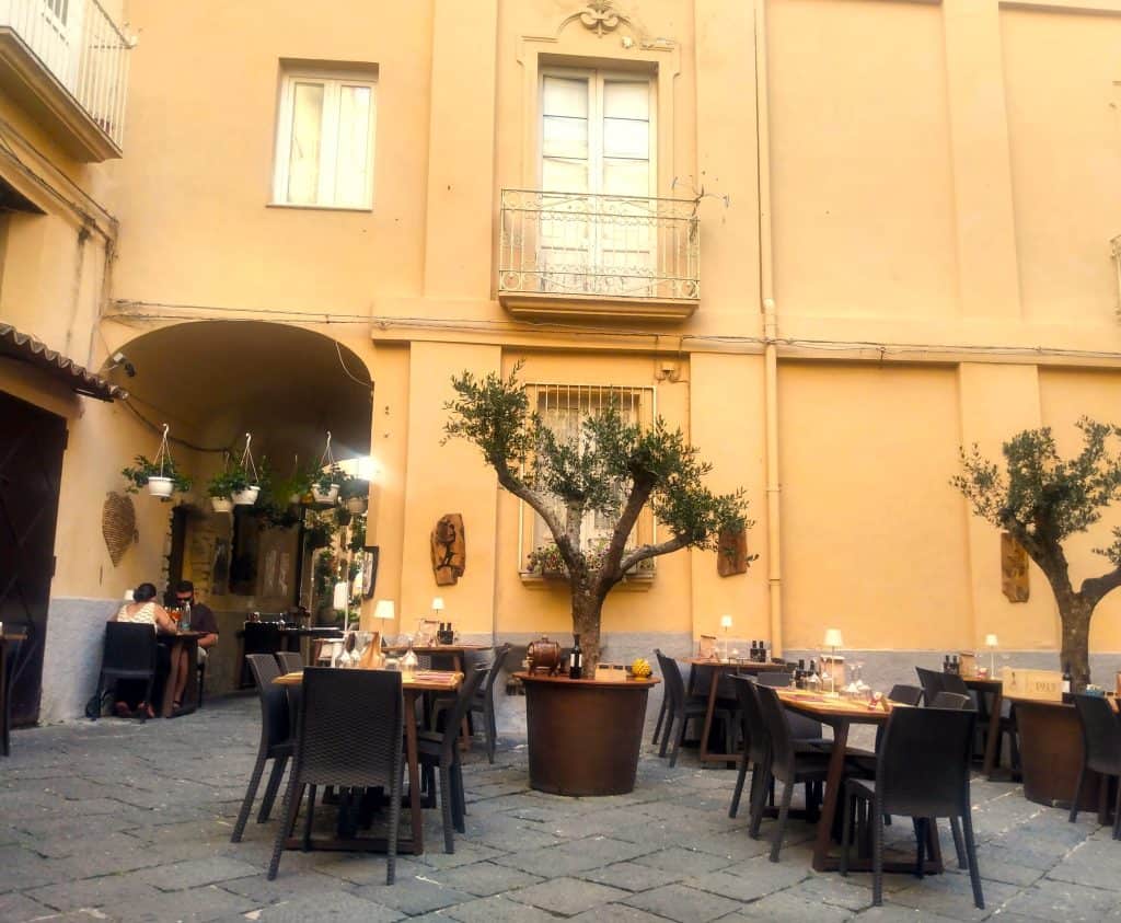 Tables and chairs in a piazza with some trees and a building in the background.